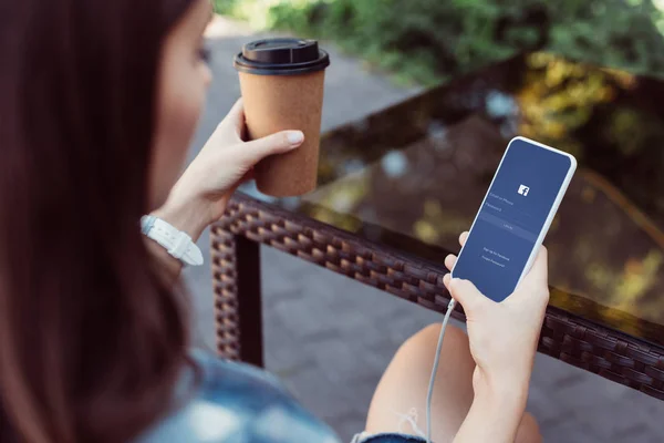 Donna che utilizza smartphone con apparecchio facebook a tavola e che tiene il caffè in tazza di carta — Foto stock