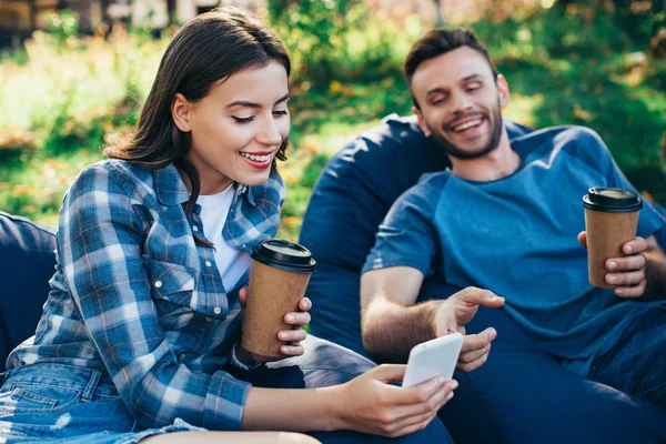 Улыбающиеся друзья смотрят на смартфон на креслах из бобов в парке и держат кофе в бумажных стаканах — стоковое фото