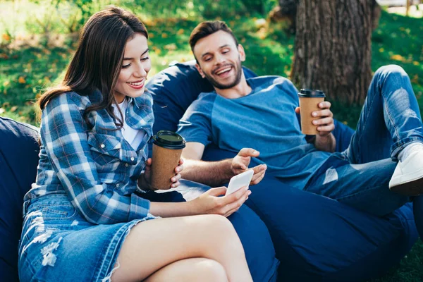 Amigos sonrientes mirando el teléfono inteligente en sillas de bolsa de frijol en el parque - foto de stock