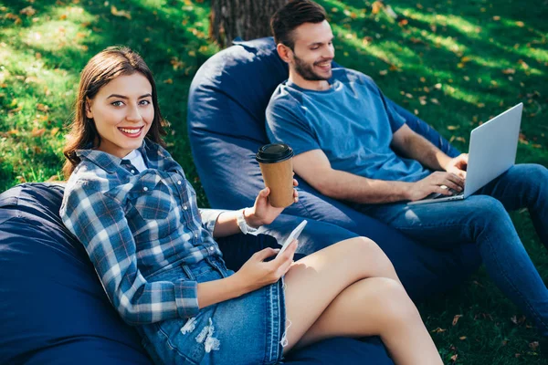 Colegas que participam de webinar sobre cadeiras de saco de feijão no parque com laptop e smartphone — Fotografia de Stock