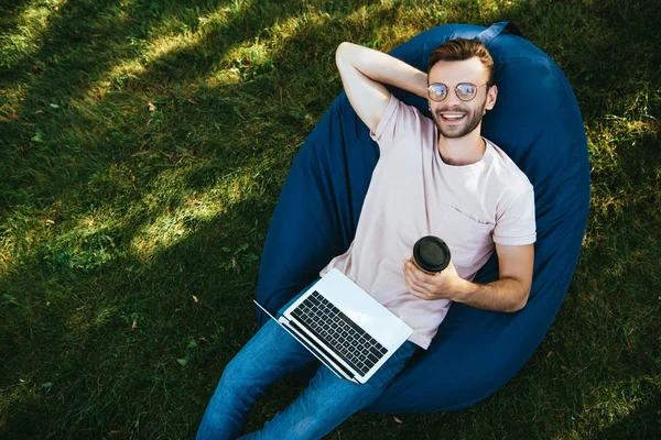 High angle view of smiling handsome man taking part in webinar and lying on bean bag chair in park — Stock Photo