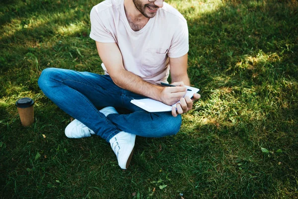 Image recadrée d'un homme souriant prenant des notes sur l'herbe verte dans le parc — Photo de stock