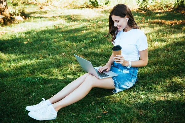 Lächelndes schönes Mädchen, das an einem Webinar mit Laptop im Park teilnimmt und Kaffee to go hält — Stockfoto
