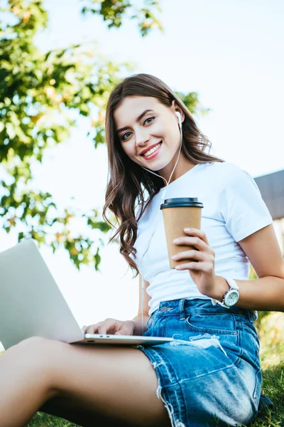 Heureuse belle fille prenant part à webinaire avec ordinateur portable dans le parc et tenant café dans une tasse en papier — Photo de stock