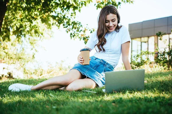 Belle fille souriante prenant part à webinaire dans le parc et tenant tasse de café jetable — Photo de stock