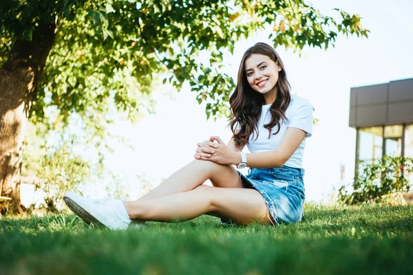 Sonriente hermosa chica sentado en verde hierba en el parque y mirando a la cámara - foto de stock