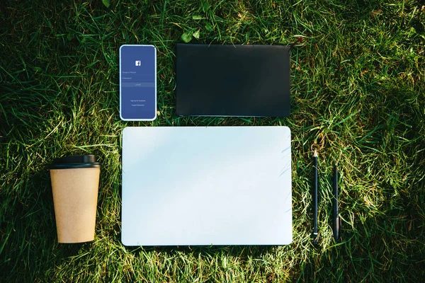 Top view of smartphone with facebook appliance and coffee in paper cup on green grass in park — Stock Photo