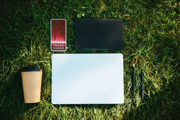 Blick von oben auf Smartphone mit Handelskursen Gerät und Kaffee im Pappbecher auf grünem Gras im Park — Stockfoto