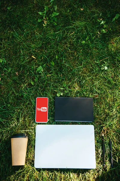 Top view of smartphone with youtube appliance, laptop and coffee in paper cup on green grass in park — Stock Photo