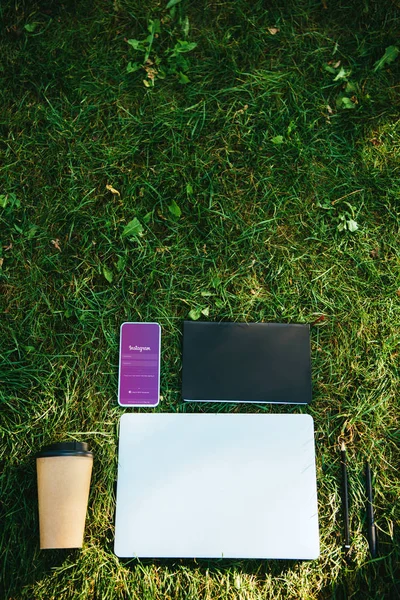 Vista superior do smartphone com instagram aparelho, laptop e café em copo de papel na grama verde no parque — Fotografia de Stock