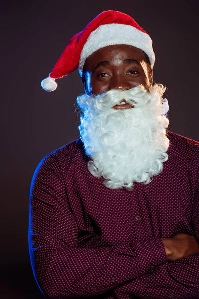 Uomo afroamericano sorridente in cappello di Babbo Natale e barba in posa con le braccia incrociate sul nero — Foto stock