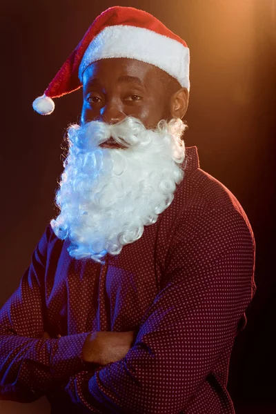 Uomo afroamericano in cappello da Babbo Natale e barba in posa con braccia incrociate su nero con retroilluminazione — Foto stock