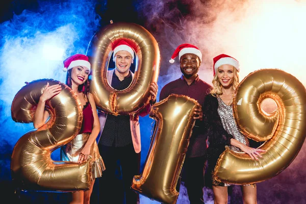 Glamorous friends in santa hats holding 2019 new year golden balloons on black with smoke and backlit — Stock Photo