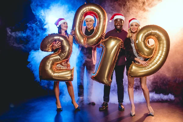 Multicultural friends in santa hats holding 2019 new year golden balloons on black with smoke and backlit — Stock Photo