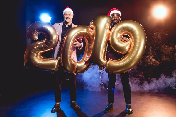 Felici amici multiculturali in cappelli di Babbo Natale che tengono 2019 nuovi palloncini d'oro anno su fumo e retroilluminazione — Foto stock