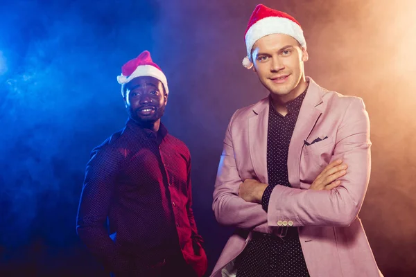 Bel homme en veste rose et santa chapeau debout près d'un ami afro-américain sur noir avec de la fumée et rétro-éclairé — Photo de stock