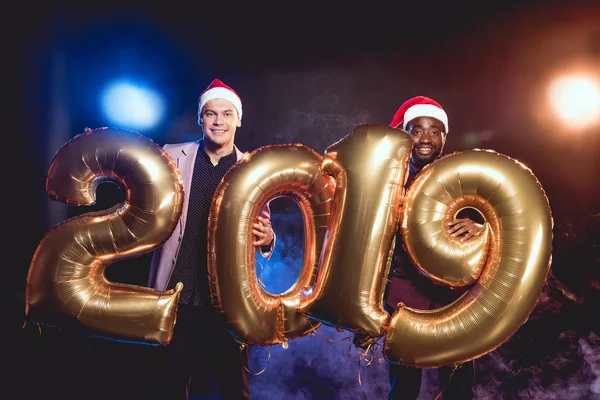 Cheerful multiethnic friends in santa hats holding 2019 new year golden balloons on smoke and backlit — Stock Photo