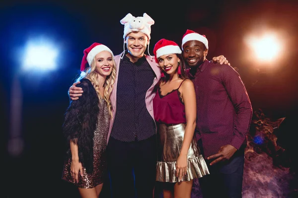 Cheerful man in pig hat posing with multicultural friends in santa hats on black with backlit — Stock Photo