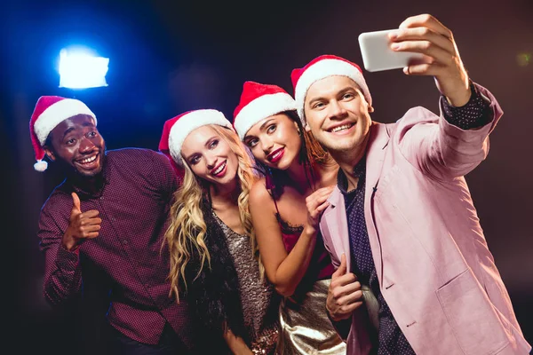 Glamorosos amigos multiculturales en sombreros de santa tomar selfie en el teléfono inteligente - foto de stock