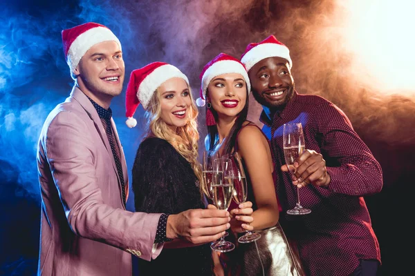 Smiling multiethnic friends in santa hats clinking with champagne glasses on new year party — Stock Photo