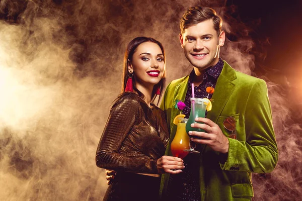 Happy glamorous boyfriend and girlfriend clinking with cocktail glasses on party with smoke — Stock Photo