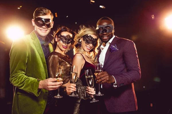 Amigos multiétnicos de lujo en máscaras de carnaval celebrando el año nuevo con copas de champán - foto de stock