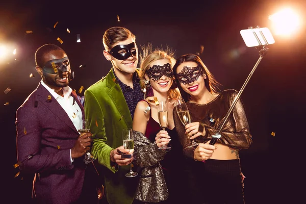 Glamorosos amigos multiculturales en máscaras de carnaval tomando selfie en el teléfono inteligente - foto de stock