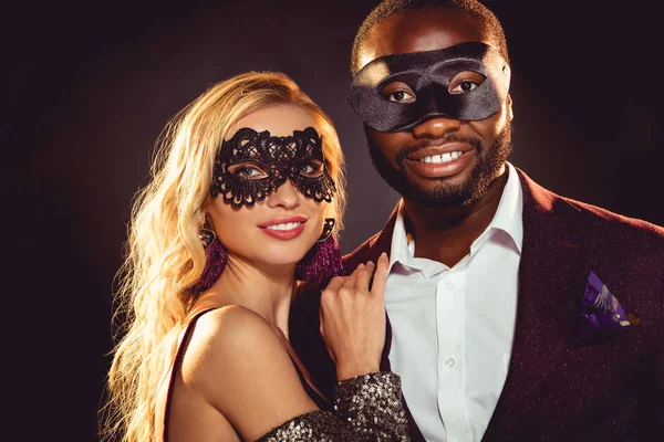 Glamouroso casal sorrindo multiétnico em máscaras de carnaval para a festa de ano novo — Fotografia de Stock