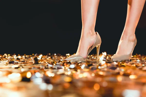 Partial view of woman in high heel shoes standing on golden confetti — Stock Photo