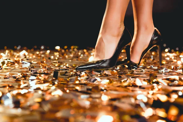 Обрезанный вид девушки в черных туфлях на высоком каблуке, стоящей на золотых конфетти — стоковое фото
