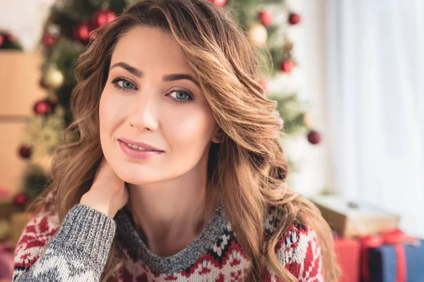Porträt einer attraktiven Frau im Winterpullover, die zu Hause am Weihnachtsbaum sitzt und in die Kamera blickt — Stockfoto