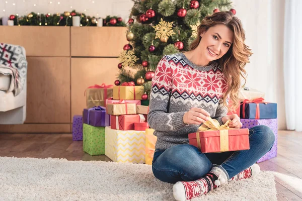 Attraente donna in maglione preparare regalo per la vigilia di Natale a casa e guardando la fotocamera — Foto stock