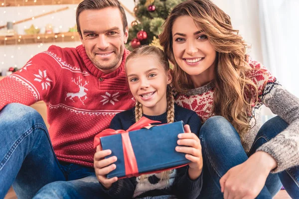 Sonrientes padres e hija con navidad presente mirando a la cámara en casa - foto de stock