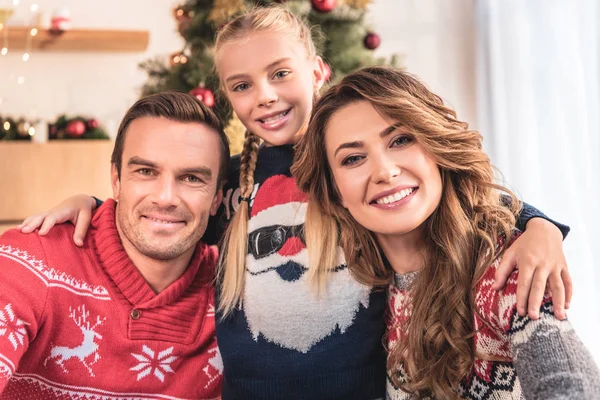 Sonrientes padres e hija en suéteres de invierno abrazando y mirando a la cámara en casa - foto de stock