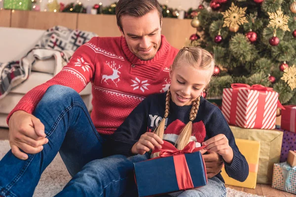 Padre abrazando hija y ella abriendo regalo cerca de árbol de Navidad en casa - foto de stock