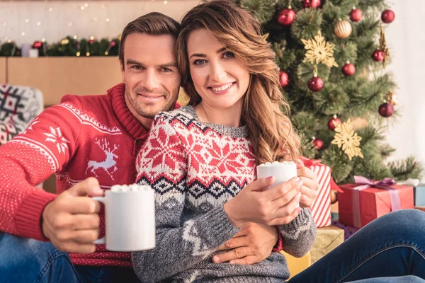 Sonrientes marido y mujer sosteniendo tazas de capuchino cerca del árbol de Navidad en casa y mirando a la cámara - foto de stock