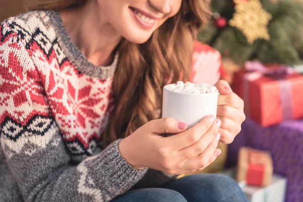 Cropped image of smiling woman holding cup of cocoa with marshmallows near christmas tree at home — Stock Photo
