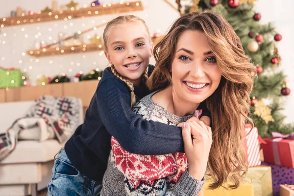 Glückliche Mutter gibt Tochter zu Hause Huckepack und sie schauen in die Kamera, Weihnachtskonzept — Stockfoto