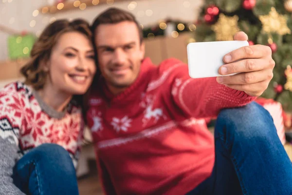 Селективный фокус улыбающейся пары в свитерах, делающей селфи со смартфоном возле рождественской елки дома — стоковое фото