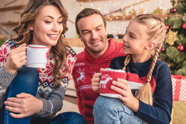 Parents gais et fille avec des tasses de Noël de cappuccino assis sur le sol à la maison — Photo de stock
