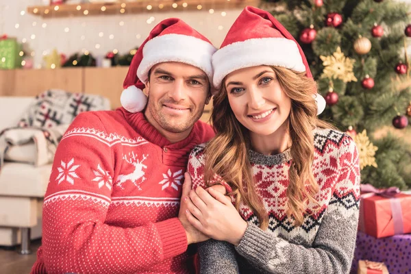 Sonrientes marido y mujer en sombreros de santa cogidos de la mano cerca del árbol de cristmas en casa, mirando a la cámara - foto de stock