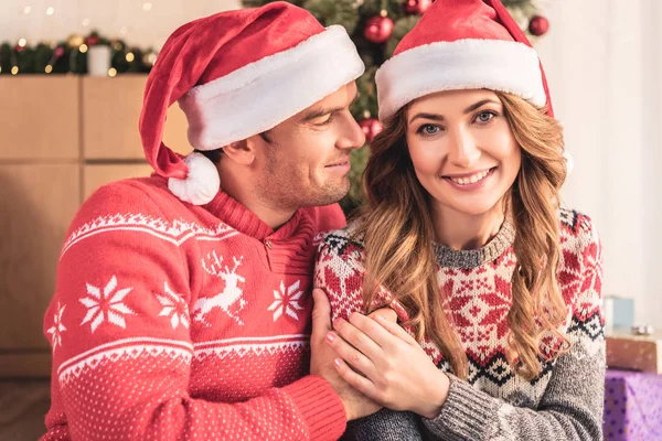 Улыбающиеся муж и жена в шляпах Санты держатся за руки возле рождественской елки дома — стоковое фото