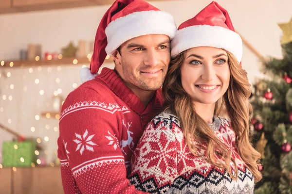 Mari souriant dans santa chapeau étreignant femme près de l'arbre de Noël à la maison et ils regardent loin — Photo de stock