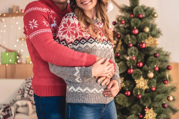 Imagen recortada de marido abrazando sonriente esposa cerca de árbol de Navidad en casa - foto de stock