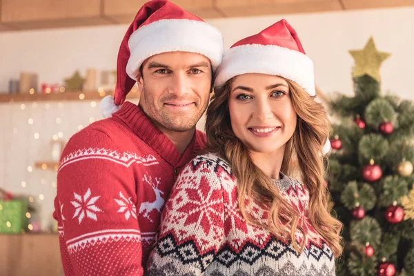 Retrato de marido y mujer en sombreros de santa abrazo cerca del árbol de Navidad en casa y mirando a la cámara - foto de stock