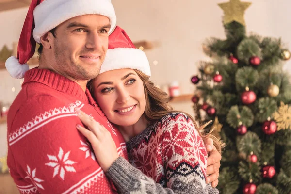 Marito e moglie in cappelli di Babbo Natale coccole vicino albero di Natale a casa e guardando altrove — Foto stock