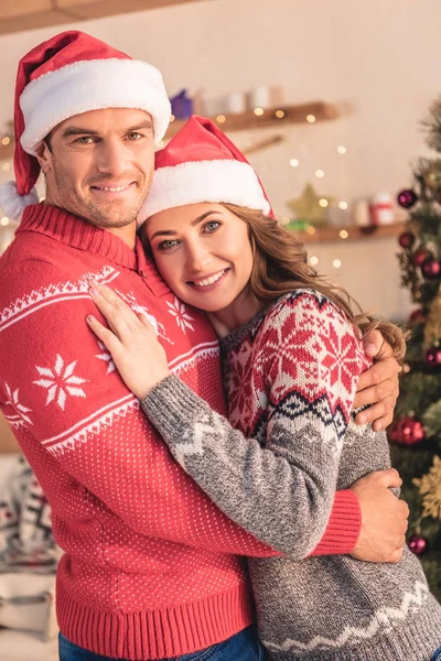 Sonrientes marido y mujer en los sombreros de santa abrazo cerca del árbol de Navidad en casa y mirando a la cámara — Stock Photo