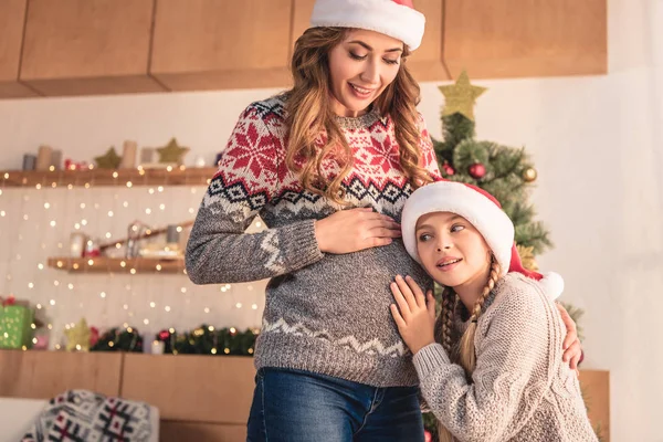Daughter in santa hat listening pregnant mother belly at home — Stock Photo