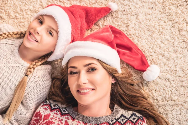 Top view of mother and daughter in santa hats lying on carpet and looking at camera at home — Stock Photo