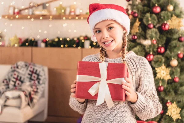 Adorable preteen kid in santa hat showing gift box and looking at camera at home — Stock Photo